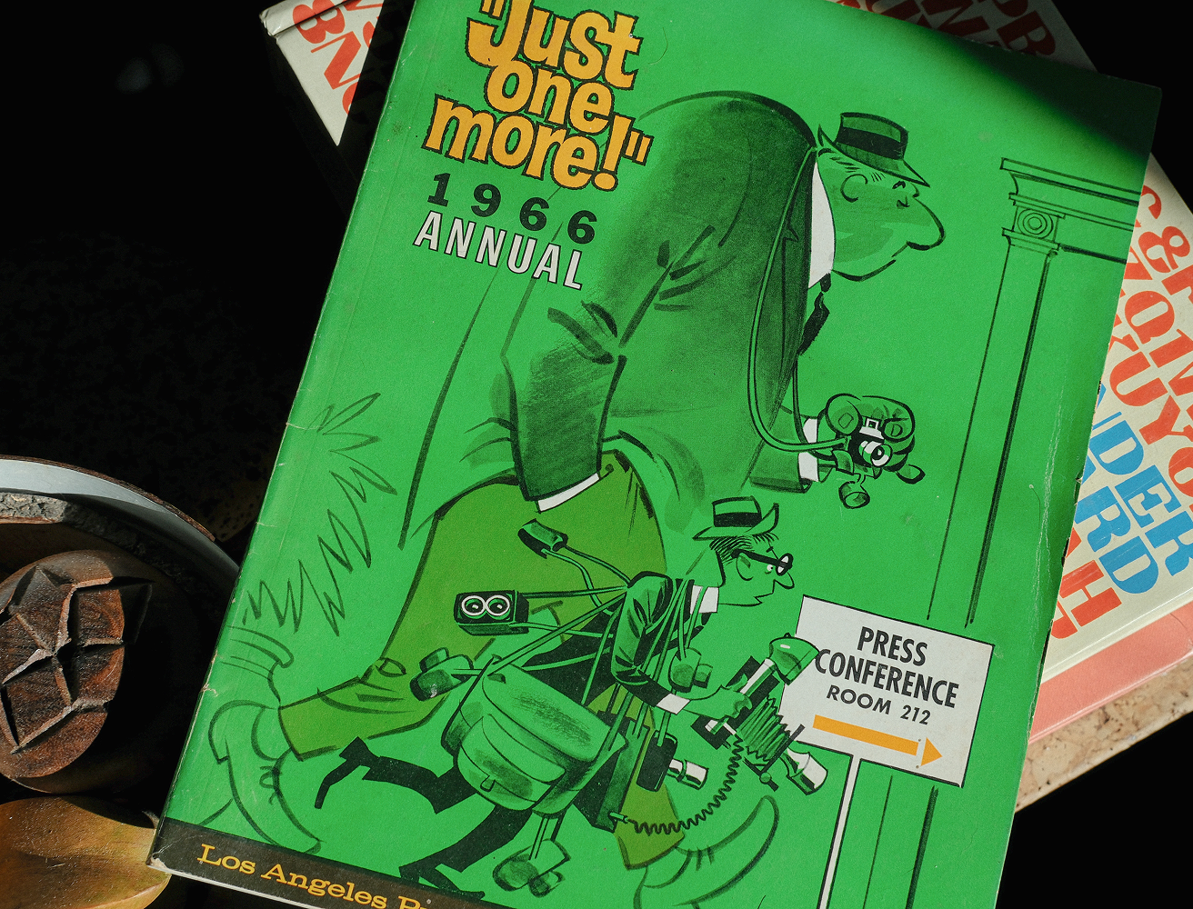 A still-life photograph showing the Just One More! 1966 Annual lying at an angle on a dark surface. The bright green cover with Karl Hubenthal’s cartoon of overburdened press photographers stands out sharply against the background. Other vintage books or magazines are partially visible beneath it, and a textured wooden object sits nearby, creating a layered, archival tableau lit with warm, directional light.